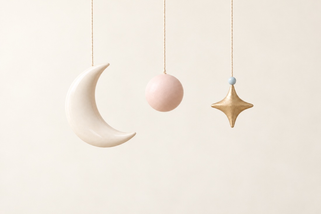 Porcelain crescent moon, blush pink sphere, and gold star with a powder-blue bead hanging against a warm cream background.